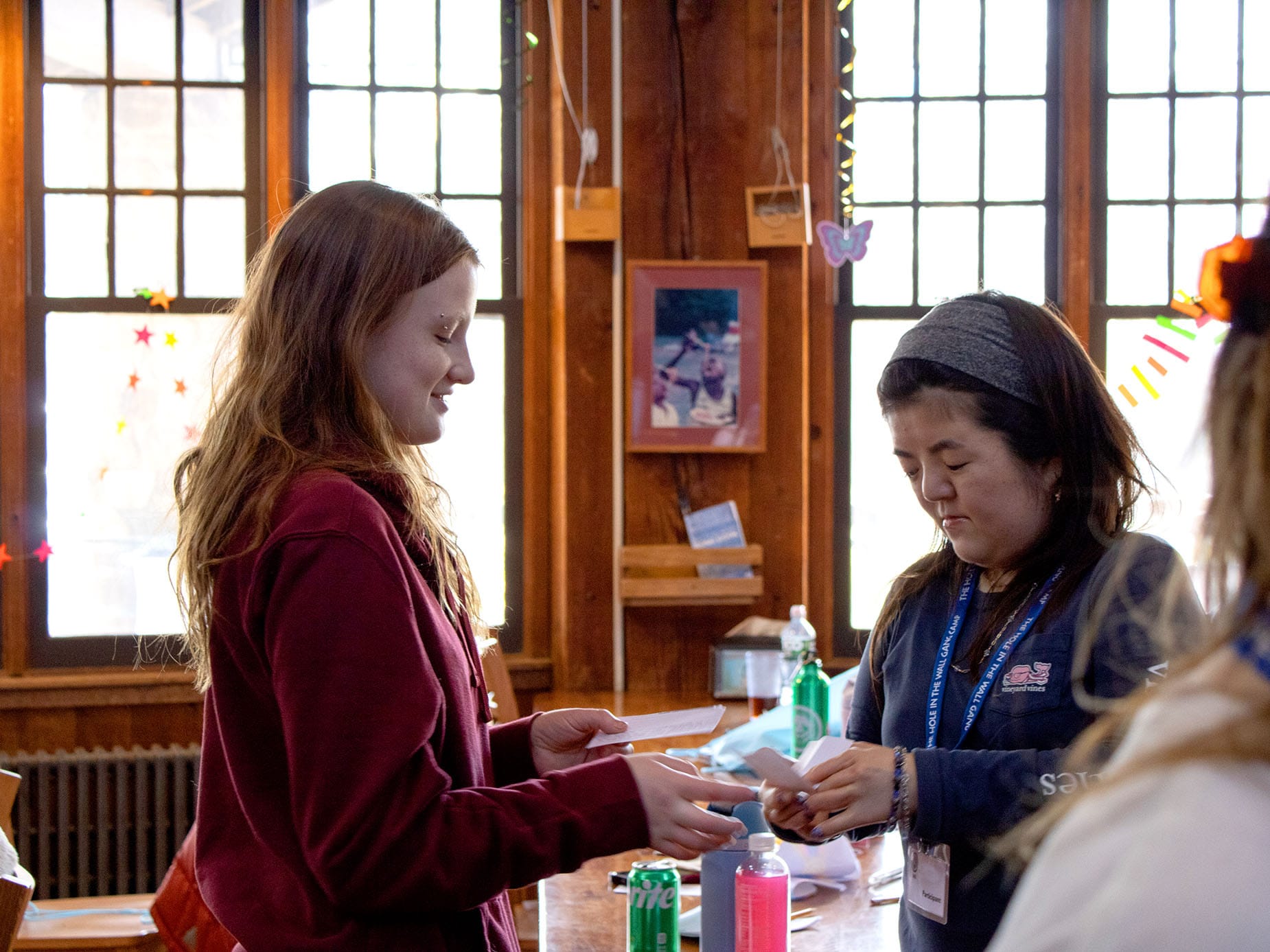 Two women stand at a wooden table indoors, one handing an object to the other. Sunlight streams through large windows behind them, and various decorations hang around the room.