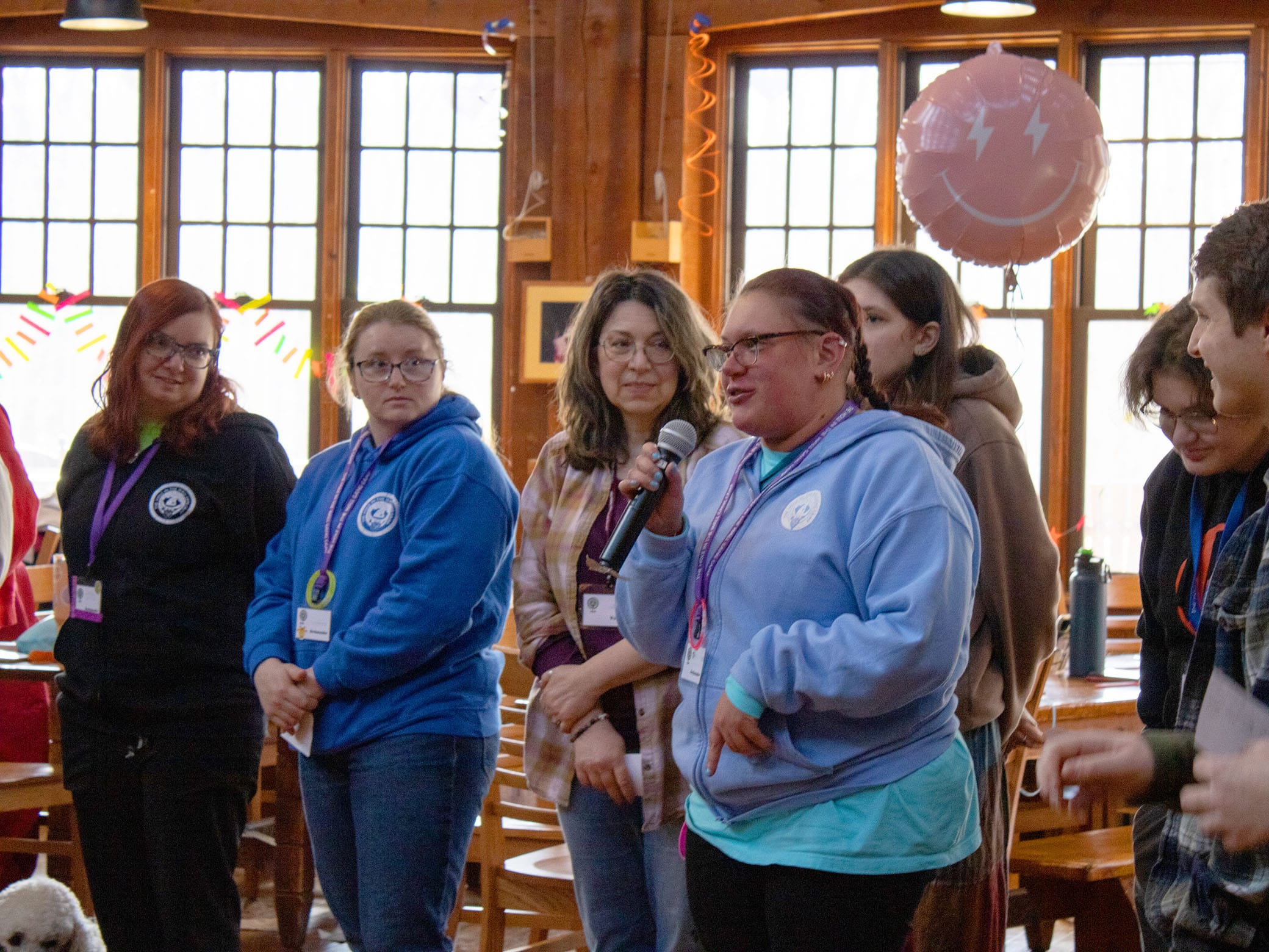 A group of people stand in a wooden room. One person in a light blue sweatshirt speaks into a microphone. Others listen and smile. A pink balloon with white designs floats in the background.