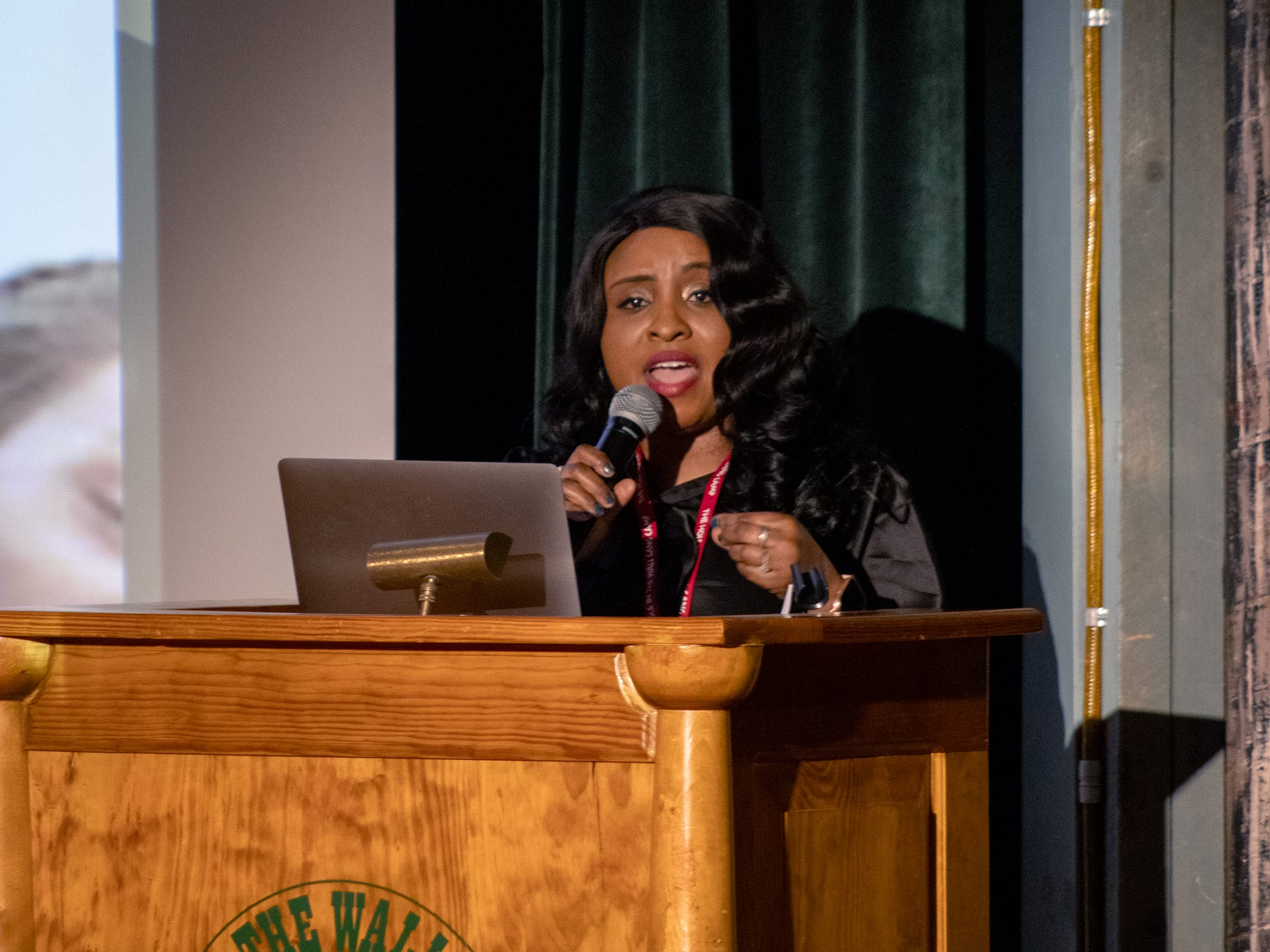 A woman speaks into a microphone while standing at a wooden podium with a laptop, in front of a dark curtain.