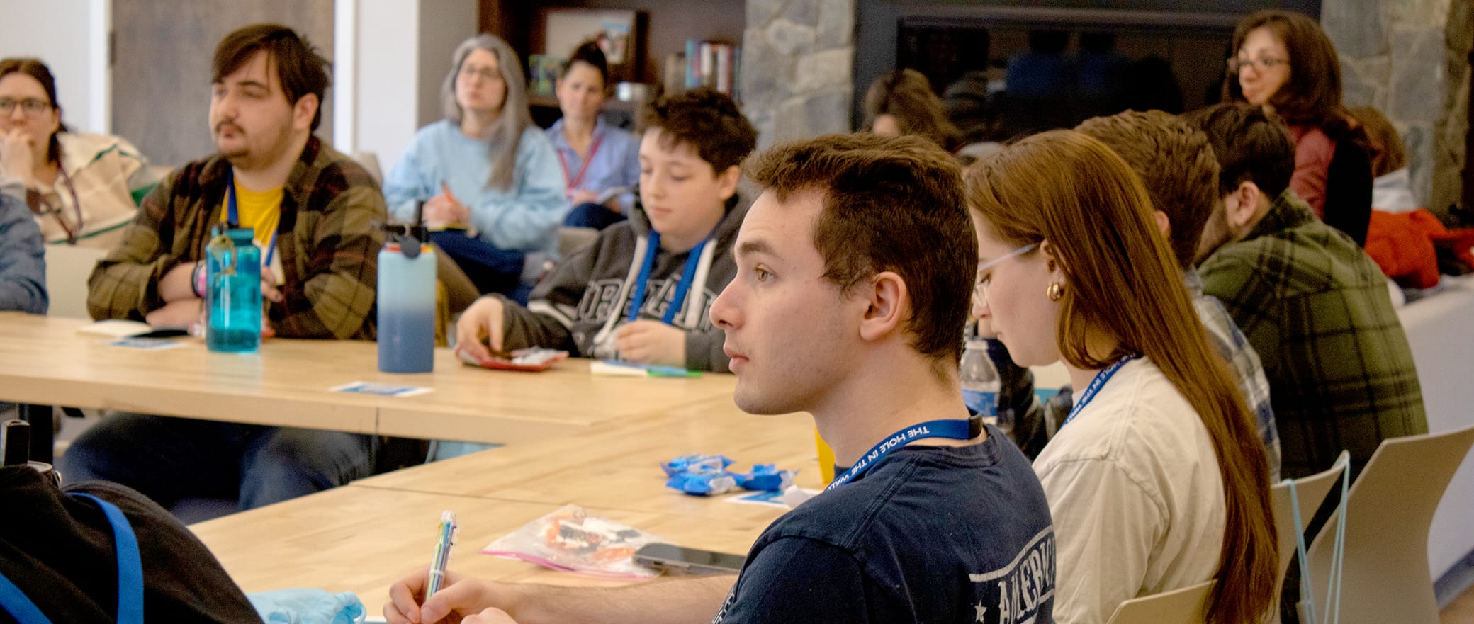 A group of people sit around tables in a well-lit room, attentively listening and taking notes during a meeting or workshop. Snacks and water bottles are on the tables.