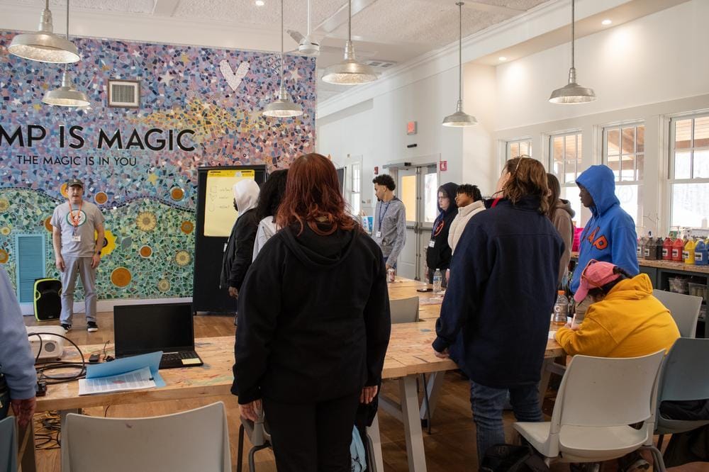 People stand and face a person speaking in front of a colorful wall that reads CAMP IS MAGIC. The room has tables, chairs, and bright lighting from large windows.