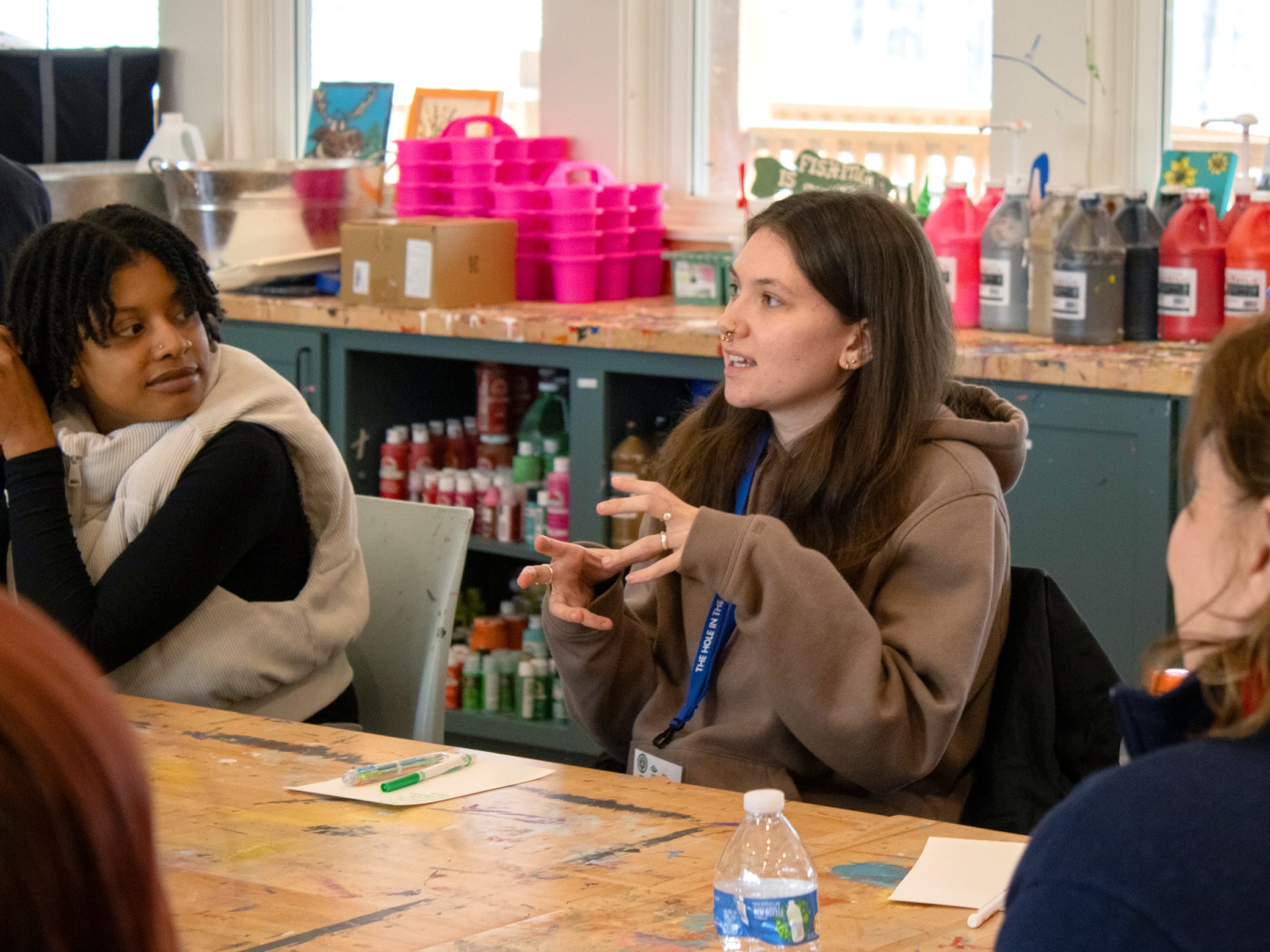 Two women sit at a paint-splattered table having a discussion. One gestures with her hands while wearing a lanyard; art supplies and paint bottles are visible in the background.