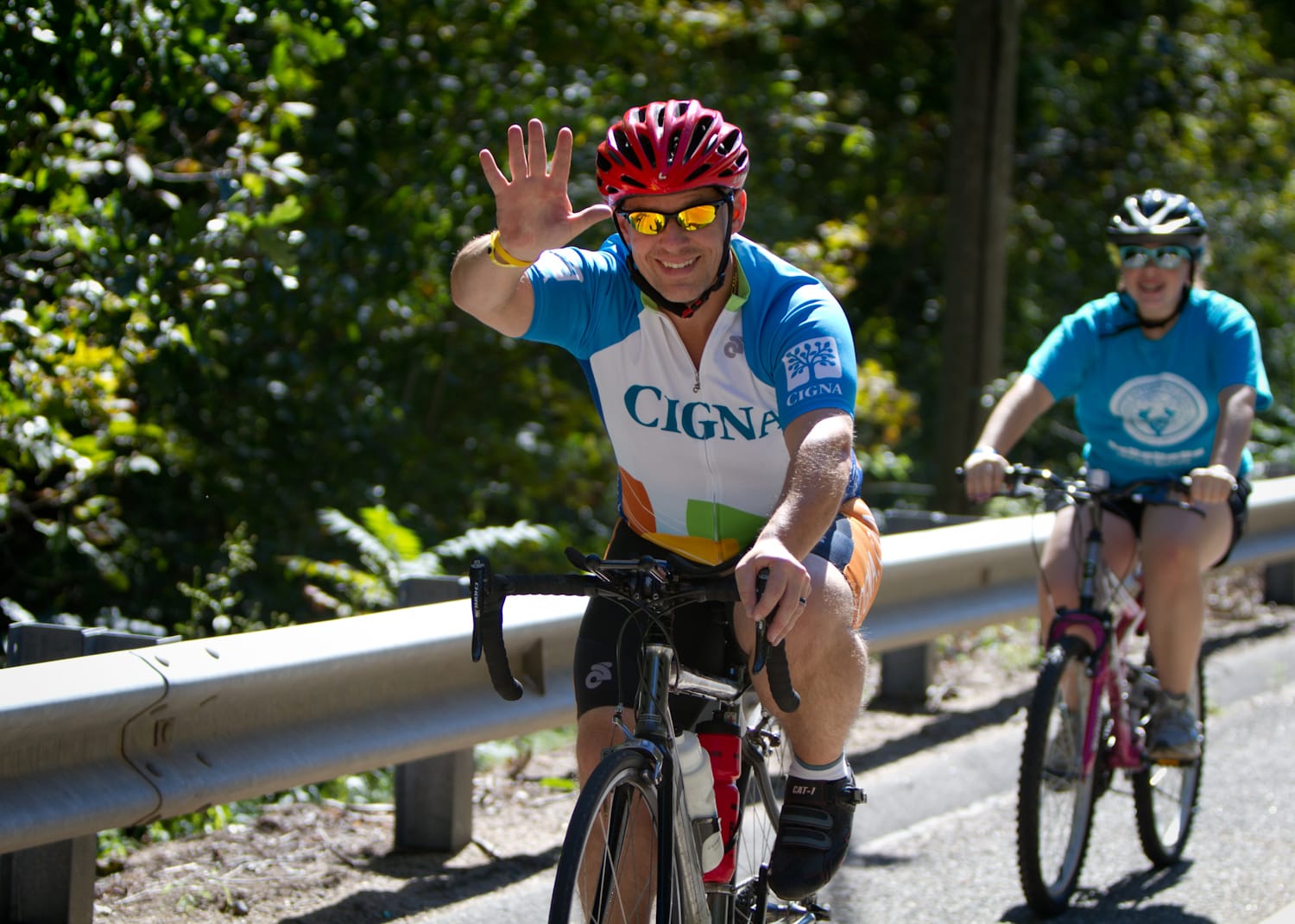 A man wearing a red helmet and sunglasses rides a bike, smiling and waving. A woman follows behind him, also biking. Both are on a tree-lined road with a metal guardrail.