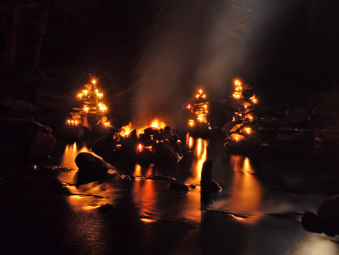 Three glowing rock cairns with small lights around a campfire are reflected on the surface of a calm stream at night, surrounded by darkness and trees, creating a warm and mystical atmosphere.