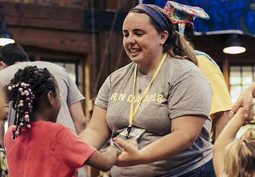 A woman in a gray Fun Day 2018 shirt smiles while holding hands and dancing with a young girl in a red shirt. Other people are visible in the background in a lively indoor setting.