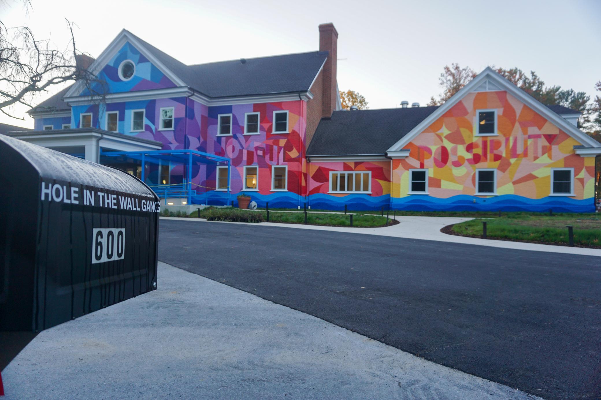 A colorful building with the words JOYFUL and POSSIBILITY painted on the side. In the foreground is a black mailbox labeled HOLE IN THE WALL GANG and the number 600.