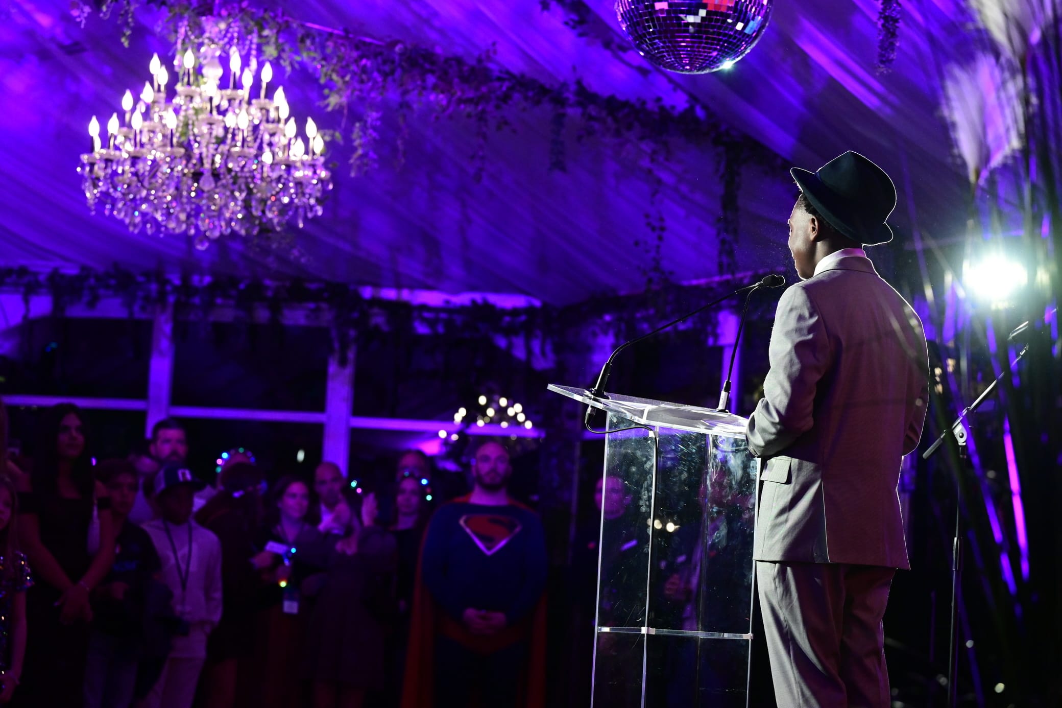 A person in a suit and hat speaks at a clear podium under a chandelier and disco ball, with a purple-lit audience, including someone dressed as Superman, watching.