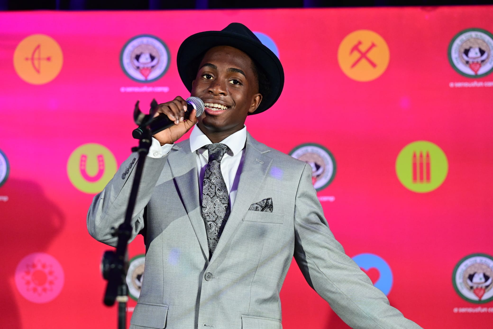 A smiling person in a gray suit, tie, and black hat sings into a microphone on stage. The background is bright pink with various colorful symbols and logos.