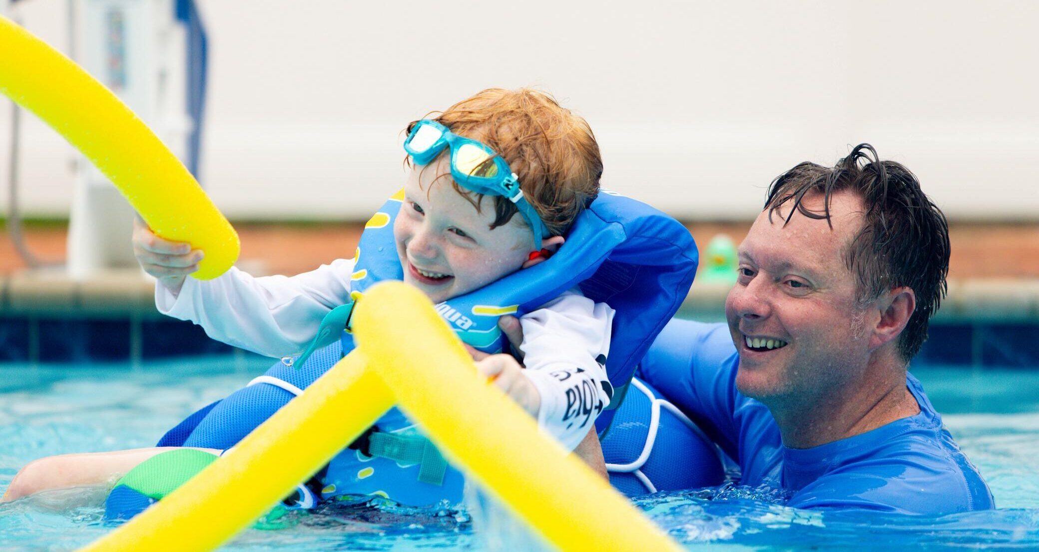 A young child wearing a life jacket and goggles smiles while playing with yellow pool noodles in a swimming pool, assisted by an adult man who is also smiling.