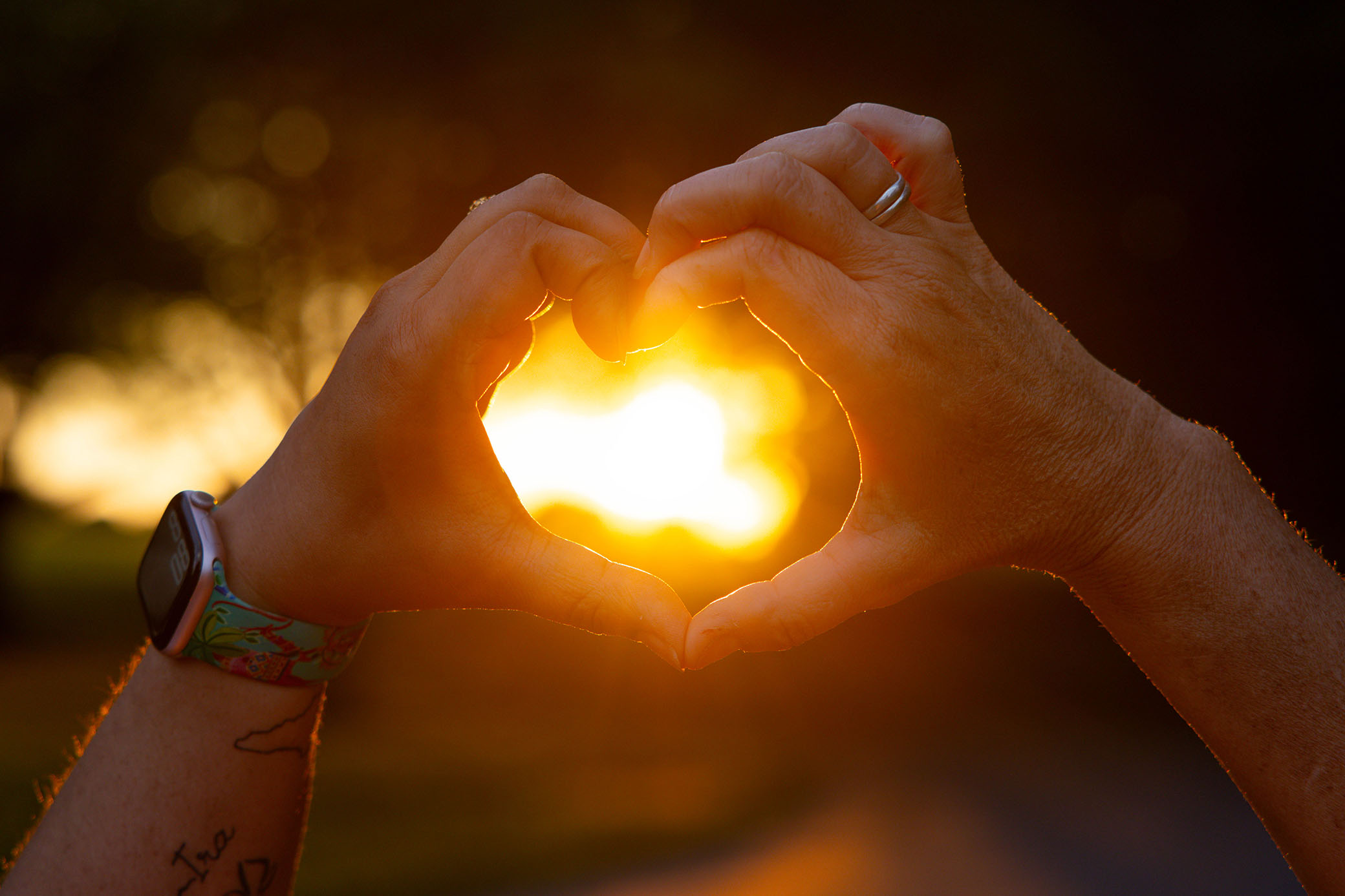 Two hands form a heart shape against a bright, golden sunset. One hand wears a colorful wristband and the other a silver ring. The background is softly blurred.