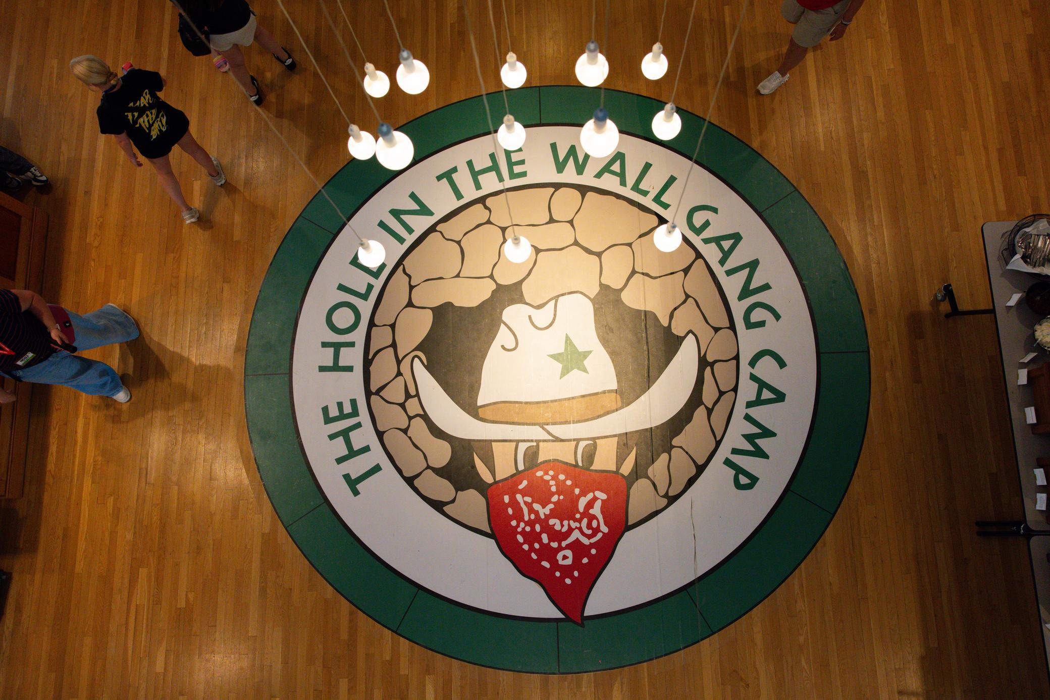 Overhead view of a circular floor logo reading The Hole in the Wall Gang Camp with a cowboy hat, red bandana, and green star, surrounded by people and hanging lights.