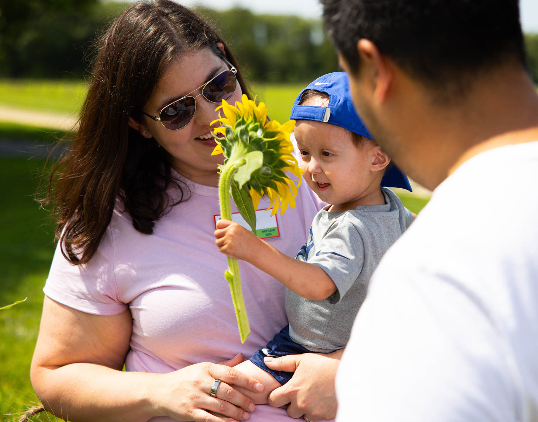 A woman holds a young child who is smiling and holding sunflowers, while an adult stands nearby. They are outdoors on a sunny day, with green grass and trees in the background.