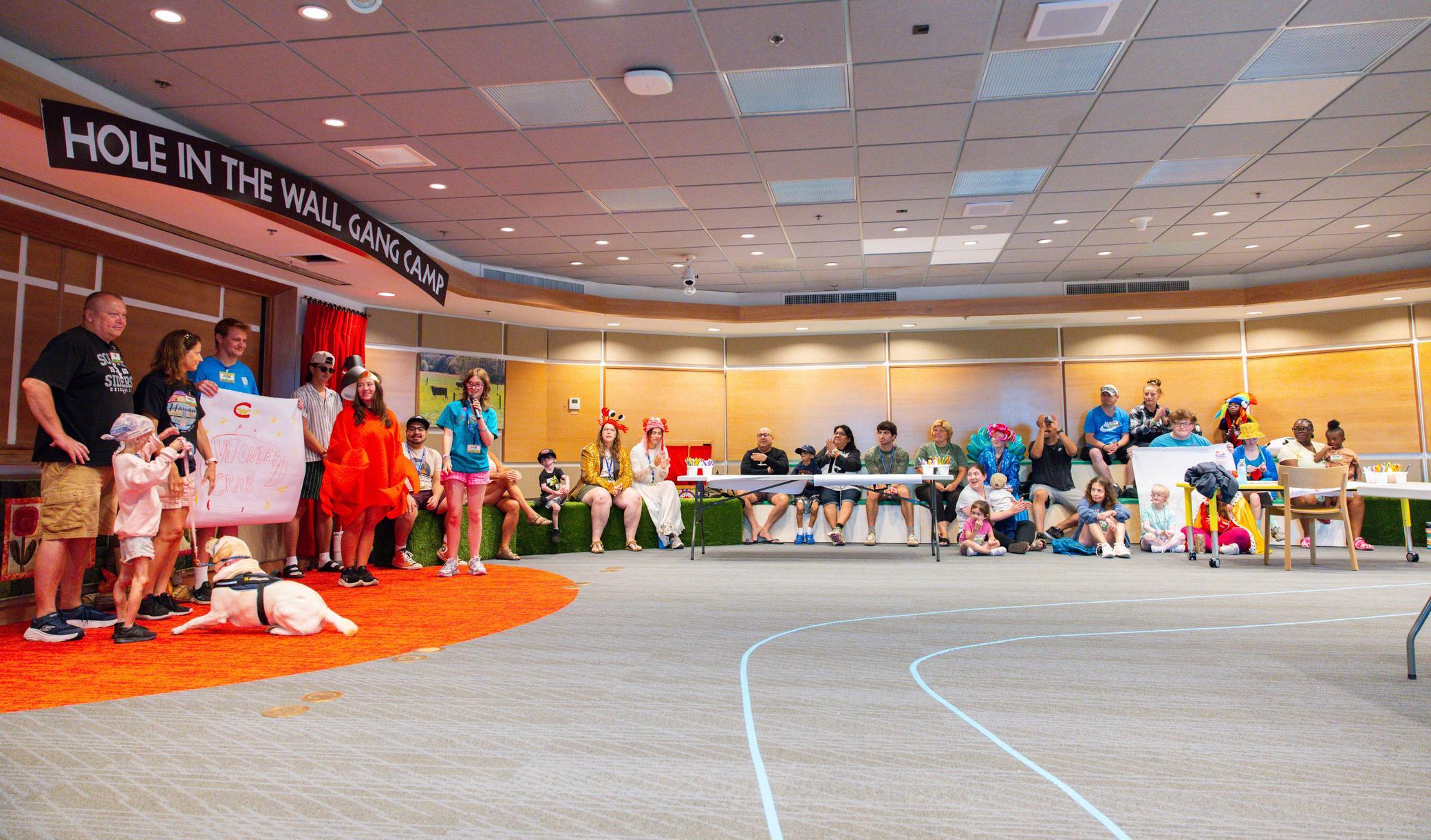 A group of people and service dogs pose in costumes on a stage, while others sit at tables watching in a large, bright room with a Hole in the Wall Gang Camp sign overhead.