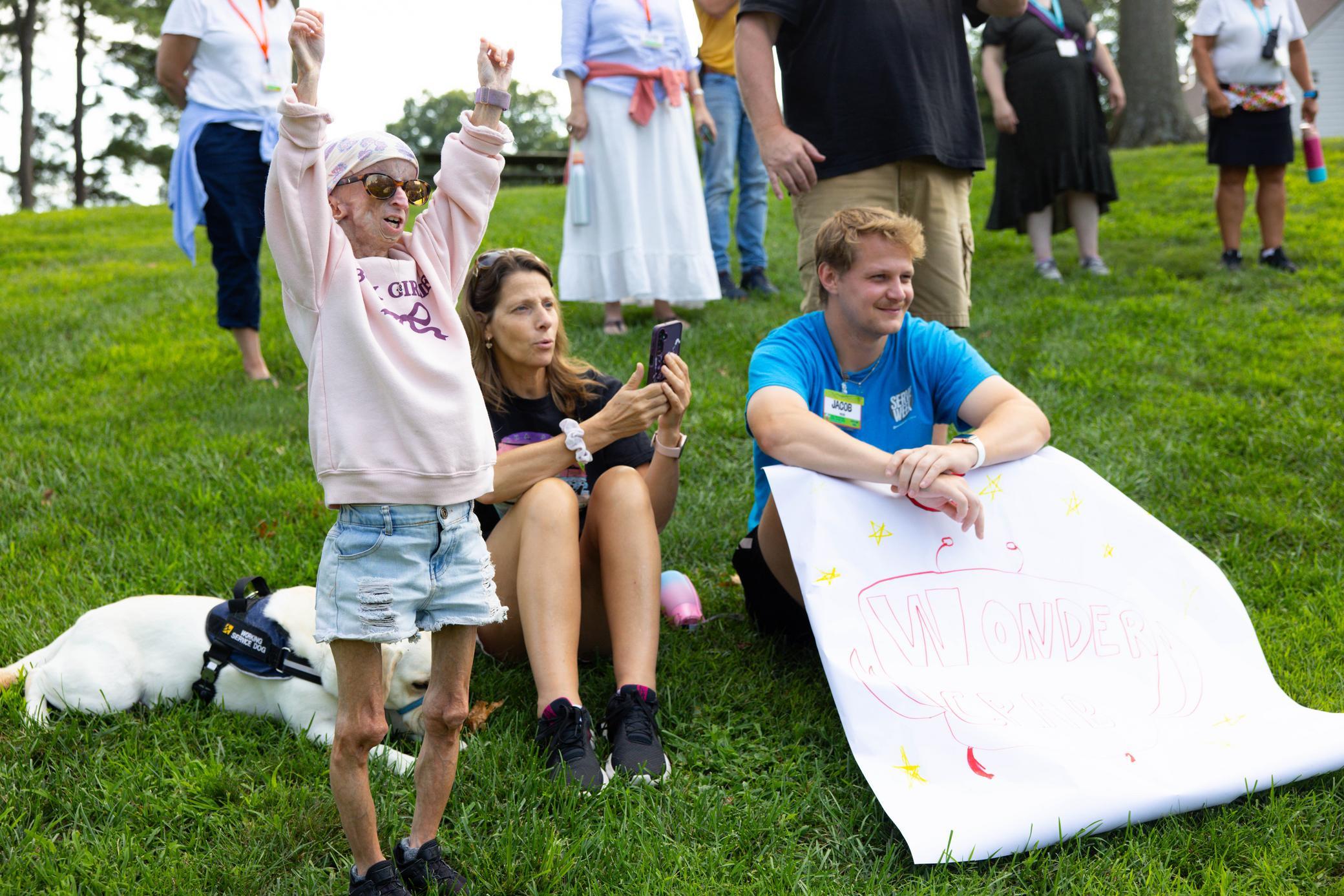 A smiling woman with arms raised stands on grass next to a seated woman using a phone and a man holding a colorful Wonders sign; other people and a dog are in the background.
