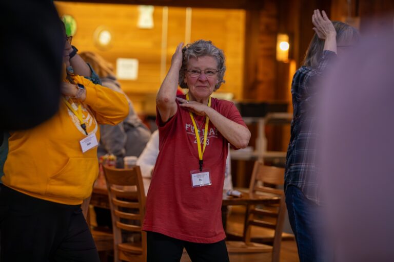 Older woman in a red shirt and name badge smiles while participating in a group activity, raising one arm and touching her head. Other people are around her in a wooden room with chairs and tables.