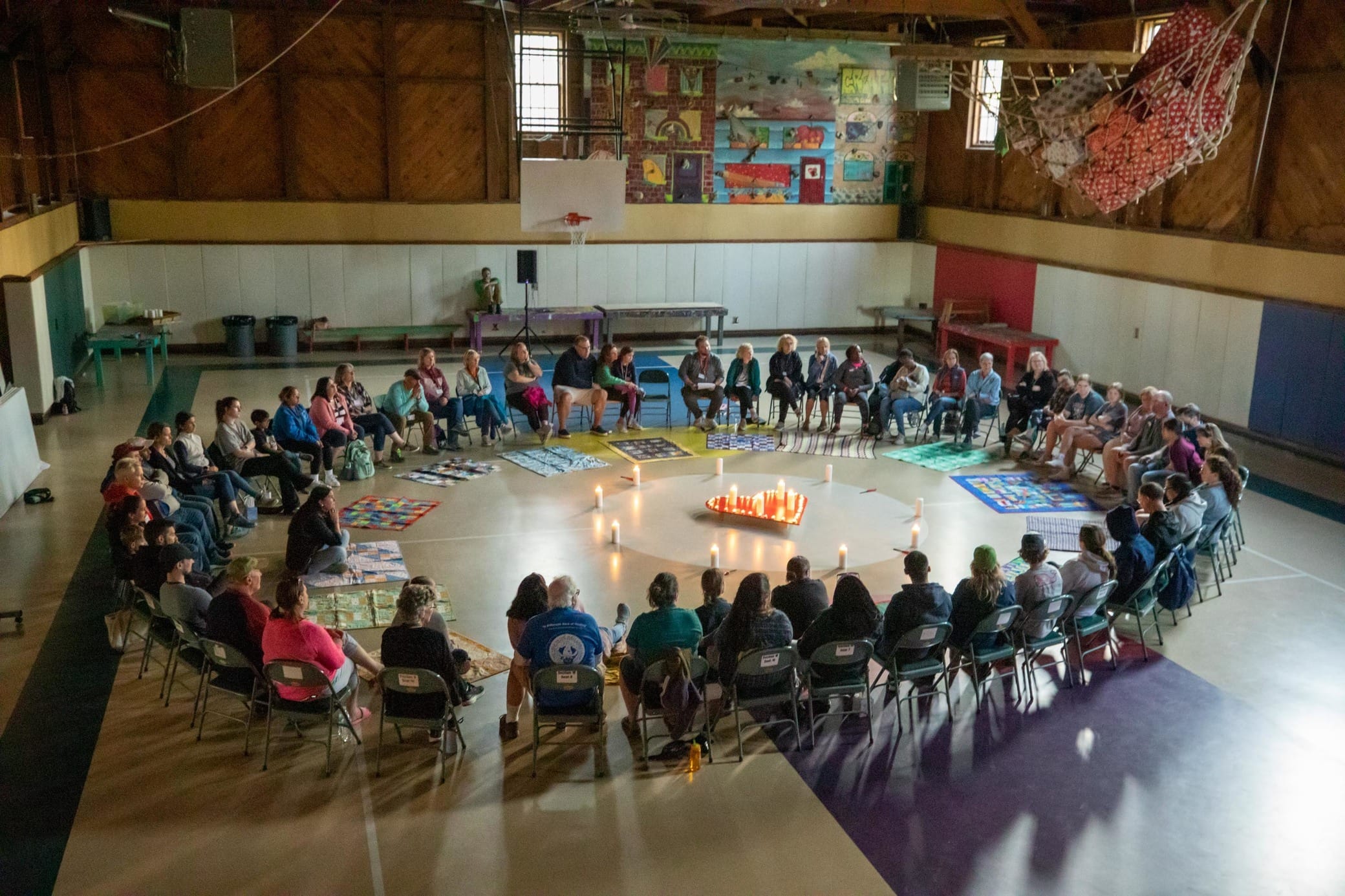 Large group of people sit in a circle inside a gym, with candles lit in the center, colorful mats on the floor, and murals on the walls in the background.