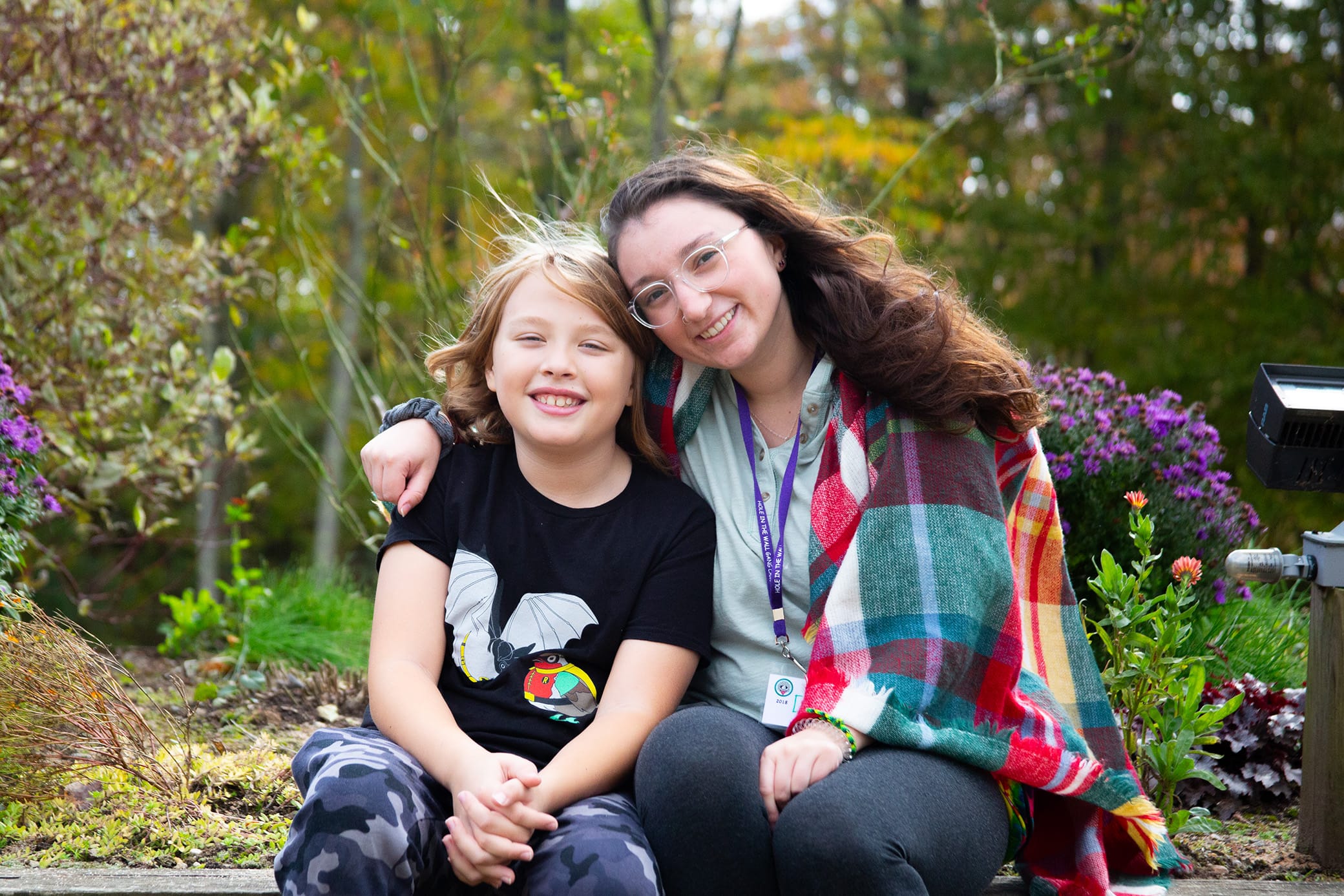 Two people sit close together outdoors, smiling at the camera. The adult has glasses and a colorful plaid shawl; the child wears a black t-shirt with a graphic. Flowers and greenery fill the background.