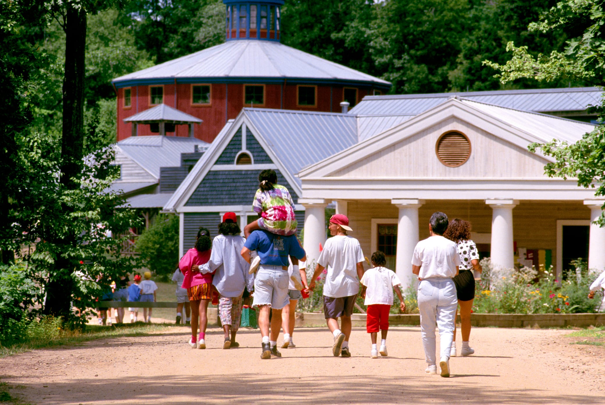 A group of people, including adults and children, walk on a dirt path toward historic-looking buildings surrounded by trees, with one child riding on an adults shoulders.