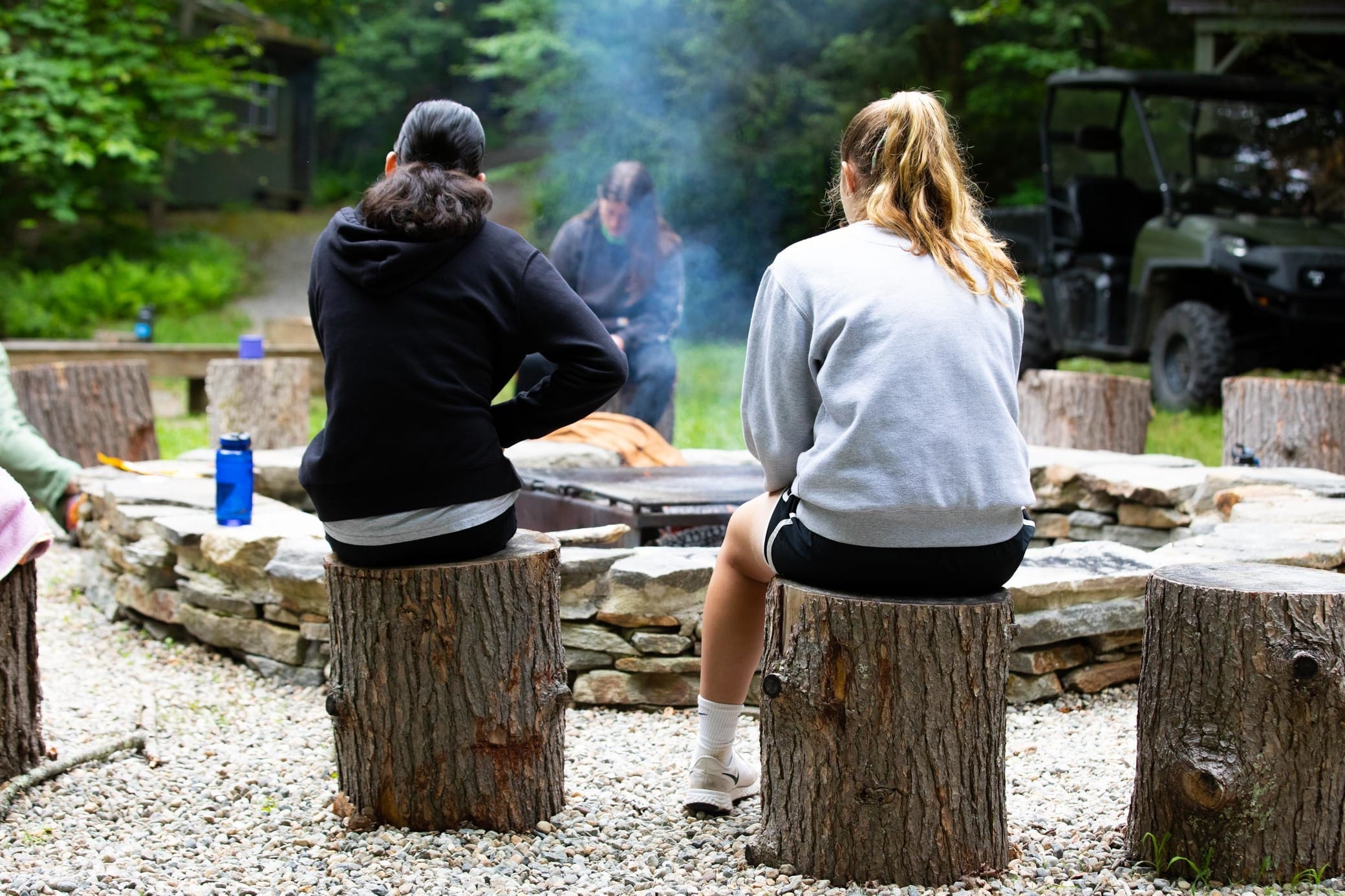 Two people sit on tree stumps around a stone campfire ring outdoors, with smoke rising and trees in the background. A third person sits by the fire, and an off-road vehicle is parked nearby.