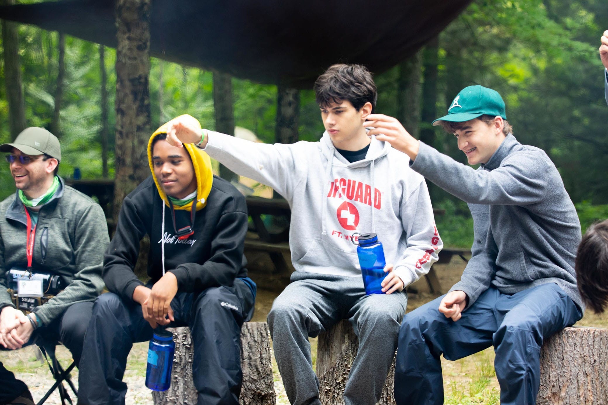 Three teens sit on logs outdoors, smiling and interacting. One wears a lifeguard hoodie and holds out a marshmallow, while the others watch and laugh. They are surrounded by trees and camping gear.