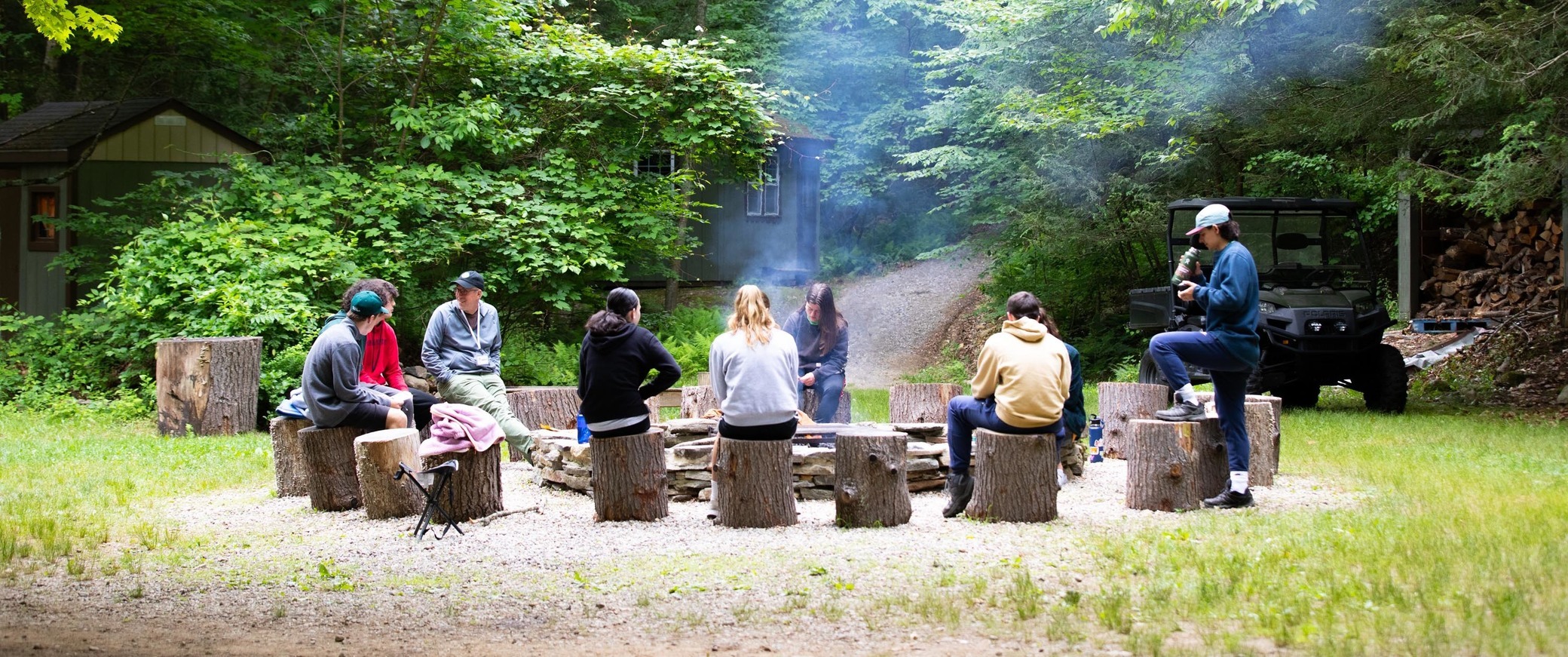 Seven people sit on tree stumps in a circle around a campfire pit in a grassy clearing surrounded by trees, with smoke rising and a small building in the background.