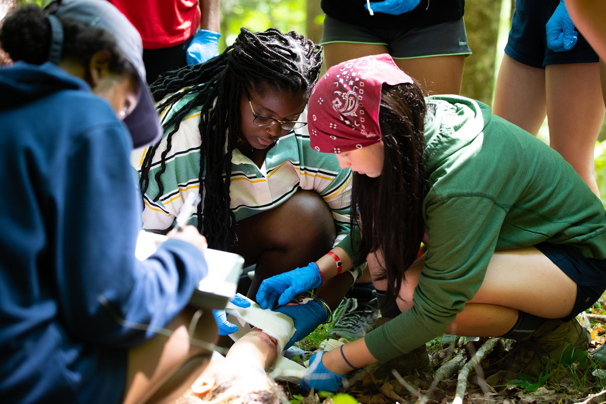 A group of young people outdoors work together to bandage a persons leg. They wear gloves and focus intently, suggesting a first aid or wilderness training scenario. Sunlight filters through the trees around them.