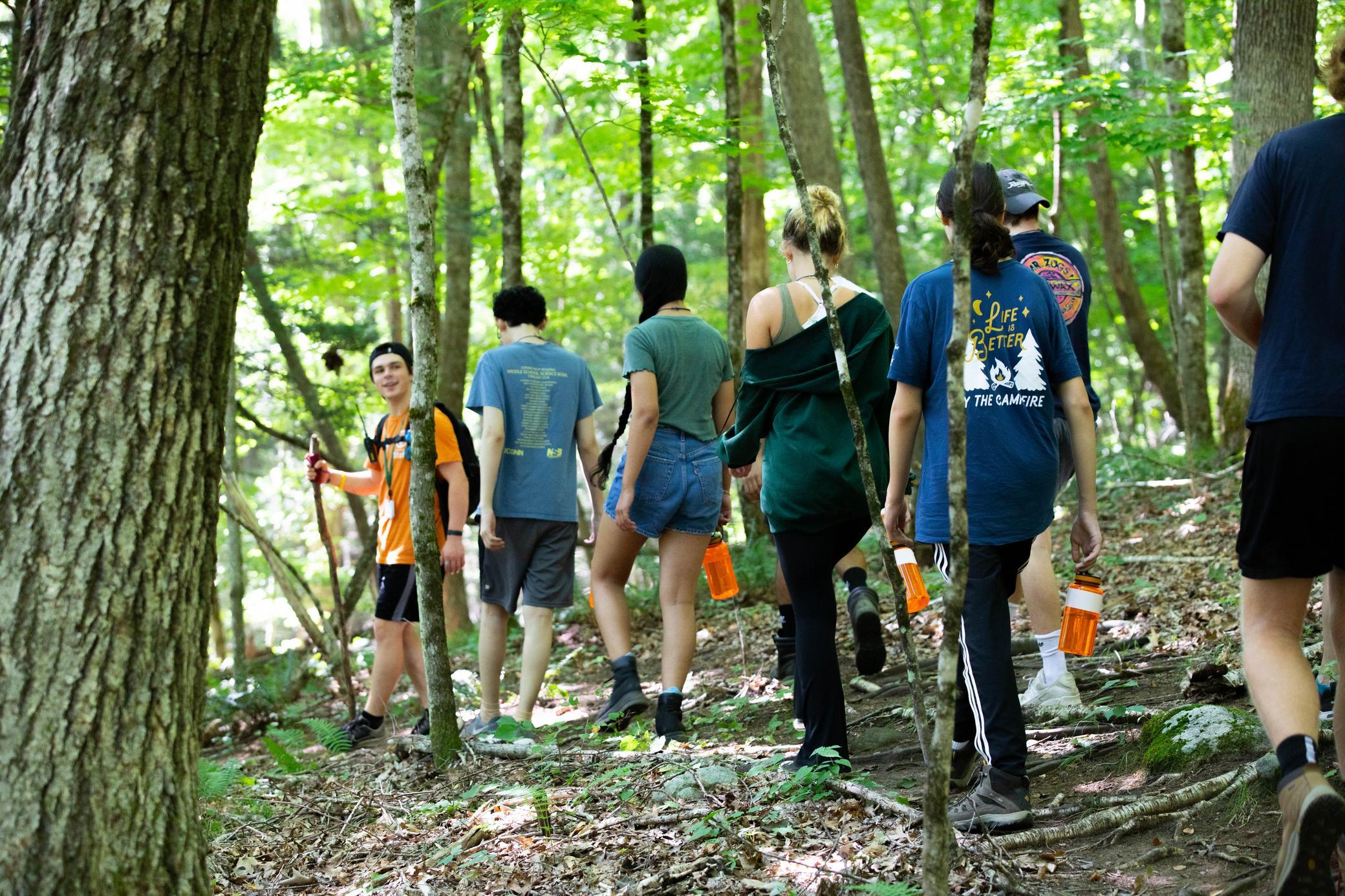 A group of people hike on a forest trail, surrounded by green trees. They wear casual outdoor clothing and carry orange water bottles. Sunlight filters through the leaves overhead.
