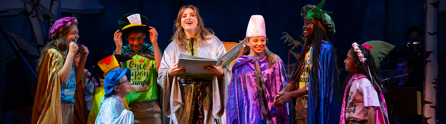 A group of children in colorful costumes stand on stage, smiling and laughing with a woman in a cape reading from a book, set against a whimsical theatrical background.