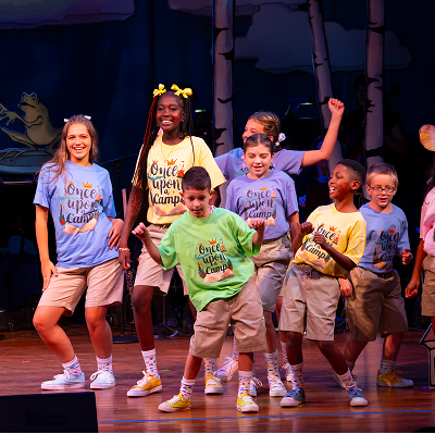 A group of children smiling and dancing on stage, wearing colorful Once Upon a Camp t-shirts and khaki shorts, performing in front of a backdrop with trees and cartoon animals.