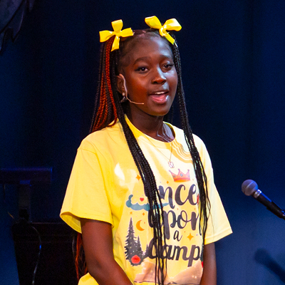 A girl with braided hair and yellow bows wears a yellow Once Upon a Camp shirt and speaks into a headset microphone on stage, with a dark blue background behind her.