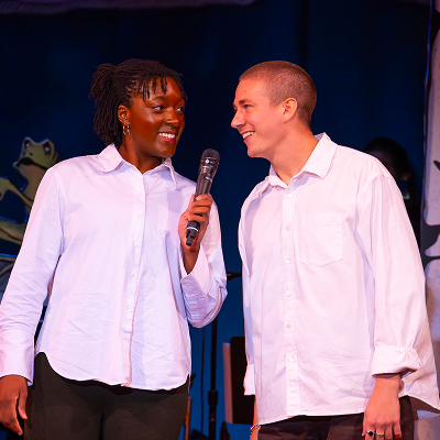 Two people in white shirts stand on stage. One holds a microphone and smiles while speaking to the other, who is also smiling. The background is dark blue with a frog illustration visible.