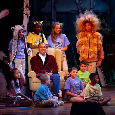 A group of children in costume and colorful shirts gathers around a man in a red robe sitting on a chair, onstage under theater lights, performing a play.