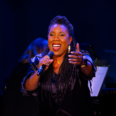 A woman with long braids sings passionately into a microphone on stage, wearing a black outfit and gold earrings, with a musician and sheet music visible in the background.
