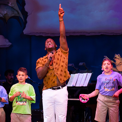 A man joyfully sings on stage, pointing upward, surrounded by children holding instruments. A piano and sheet music are visible in the background.