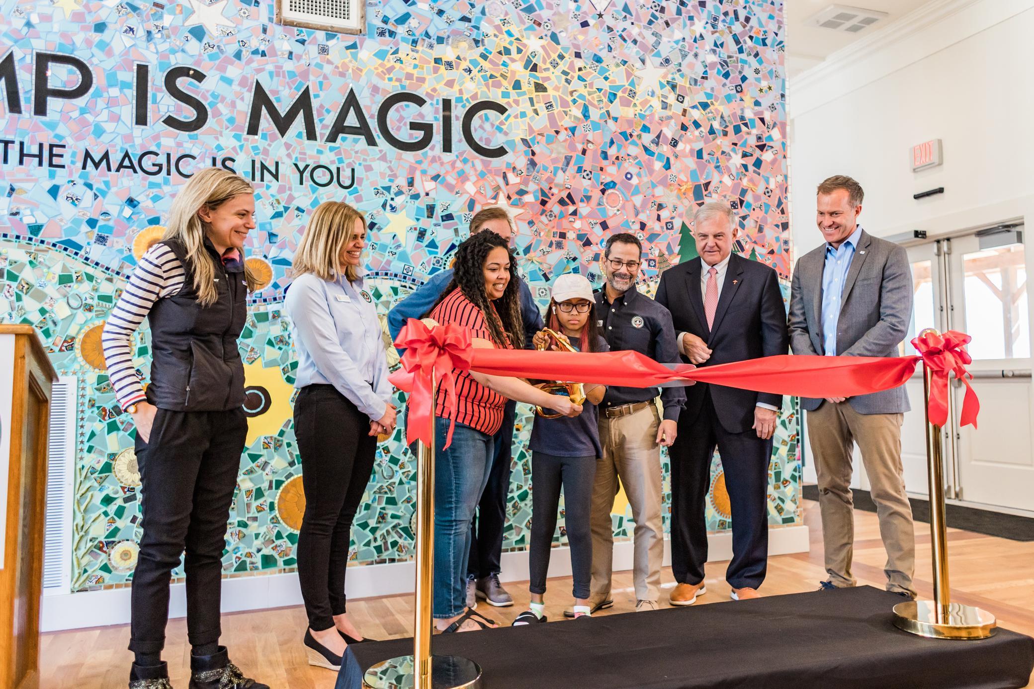 Seven people stand in front of a colorful mosaic wall, cutting a red ribbon at an indoor ceremony. The wall says, Camp is Magic. Everyone appears happy and celebratory.