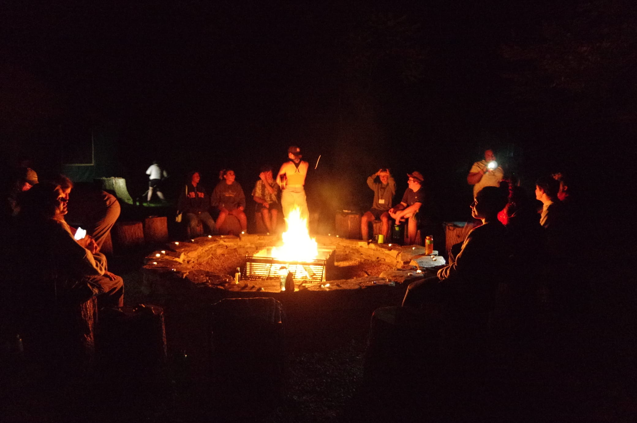 A group of people sit around a campfire at night, some holding flashlights or phones. The fire is bright, illuminating the faces and casting shadows while people relax and talk.