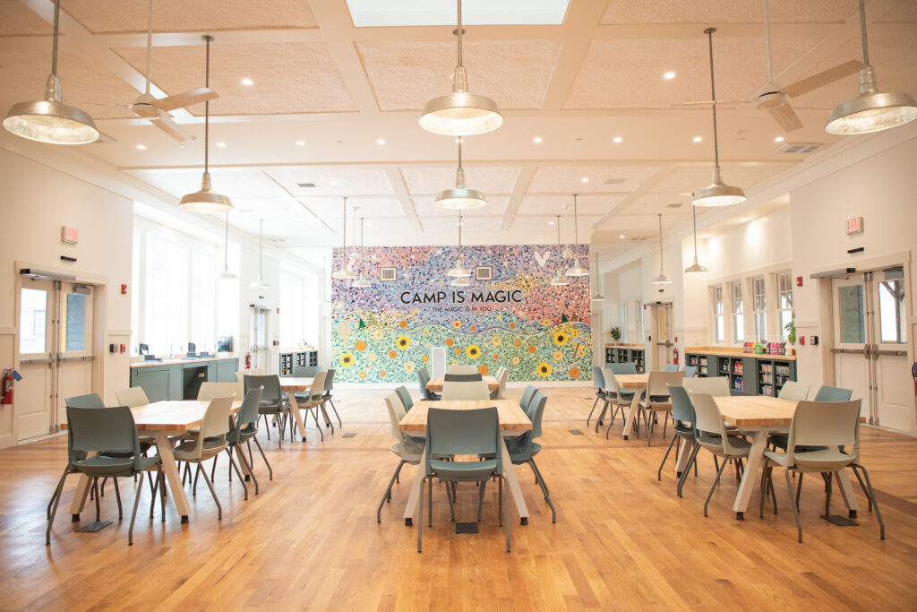A bright, spacious room with wooden floors, several tables and chairs, and a colorful mural reading CAMP IS MAGIC on the back wall. Large windows and ceiling lights add to the airy, welcoming atmosphere.