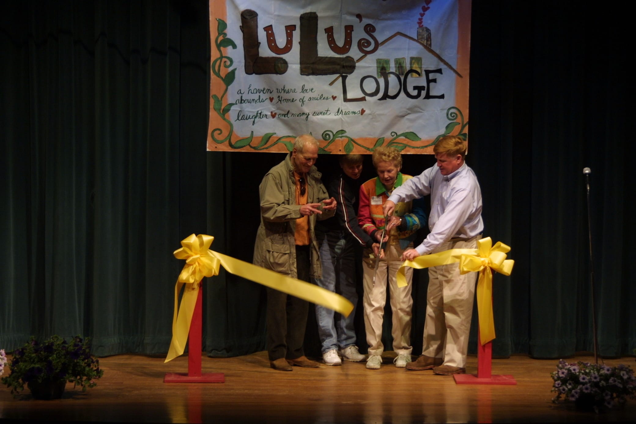 Four people cut a large yellow ribbon onstage under a LuLus Lodge banner. The stage has a dark curtain and flowers. The group appears focused and celebratory during the ribbon-cutting ceremony.