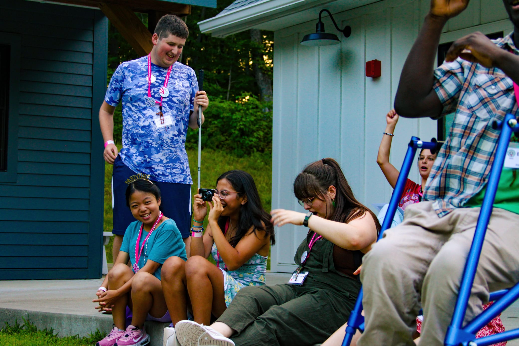 A group of young people sit and laugh outdoors by a building, some wearing name tags and wristbands, enjoying each others company during a casual gathering.