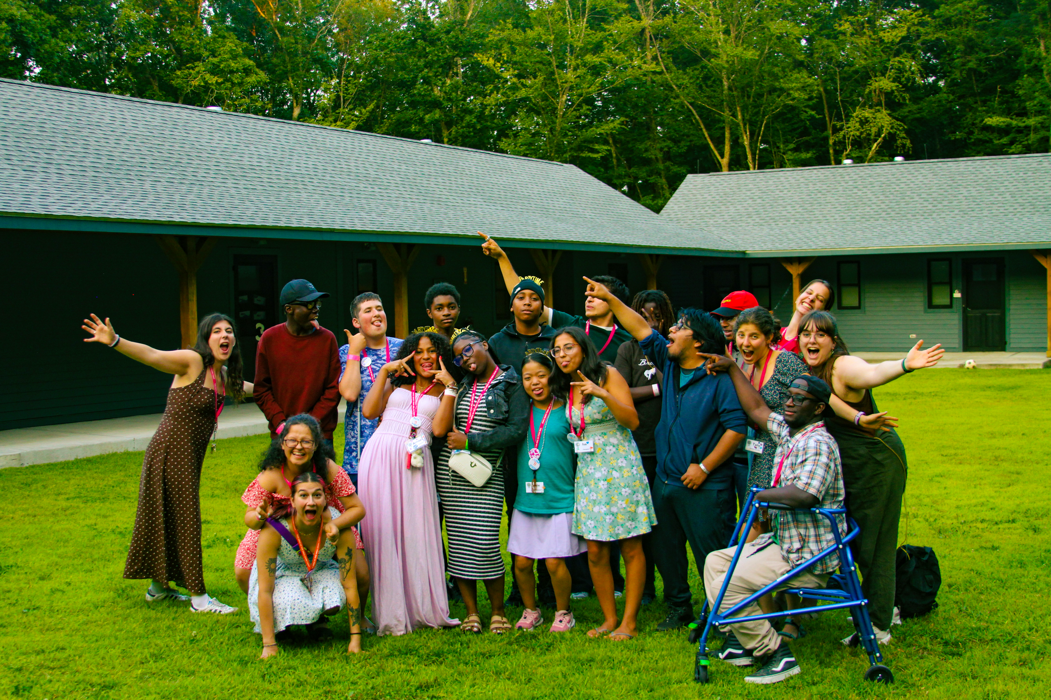 A diverse group of people, some wearing medals, smile and pose enthusiastically outside buildings with green siding. One person uses a walker. Trees are visible in the background.
