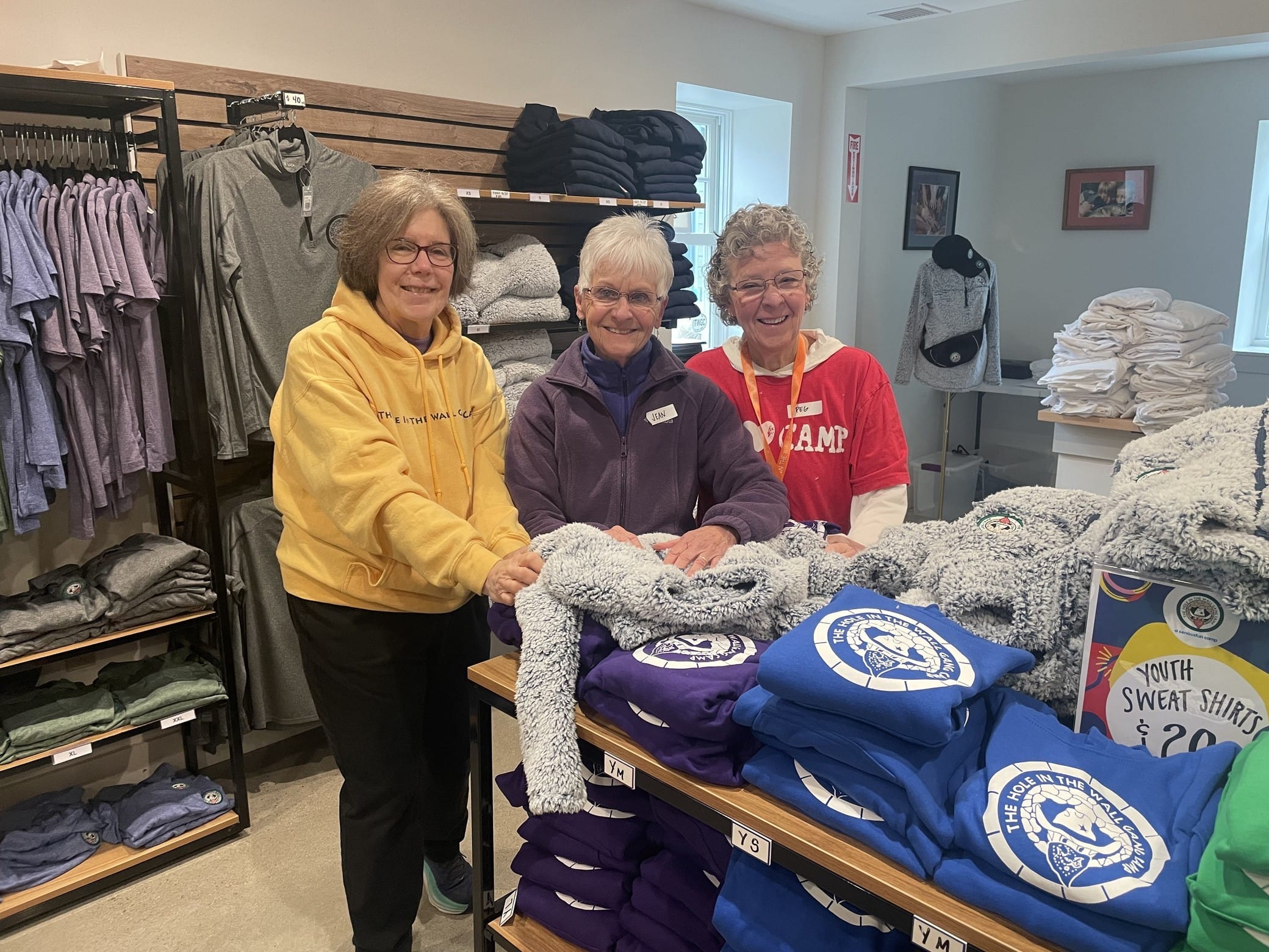 Three women smile while folding clothes in a store, surrounded by stacks of sweatshirts, t-shirts, and shelves filled with folded apparel.