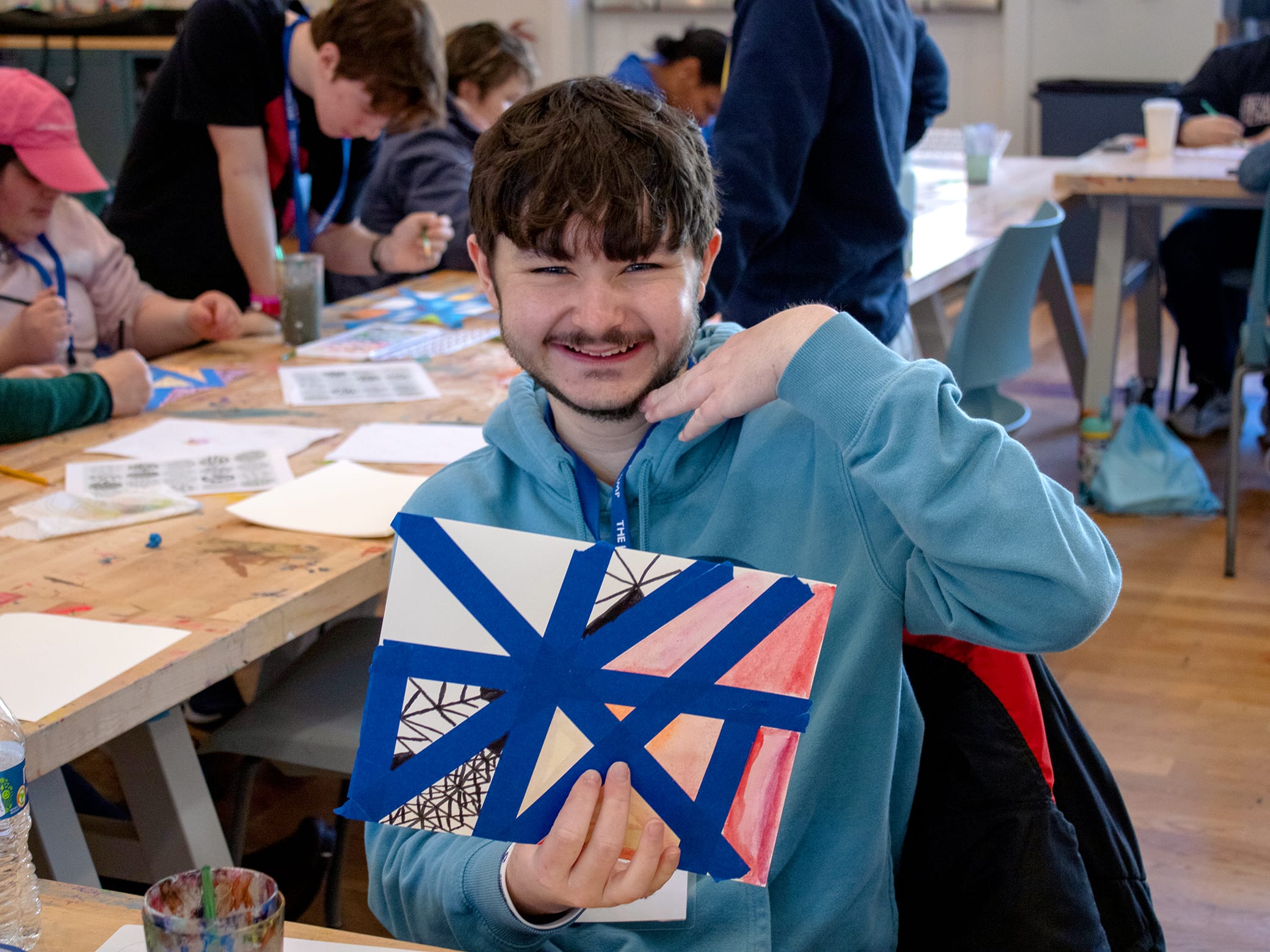 A smiling young person in a blue hoodie holds up an abstract artwork with blue tape and red, black, and white patterns, sitting at a busy art table with others in the background.