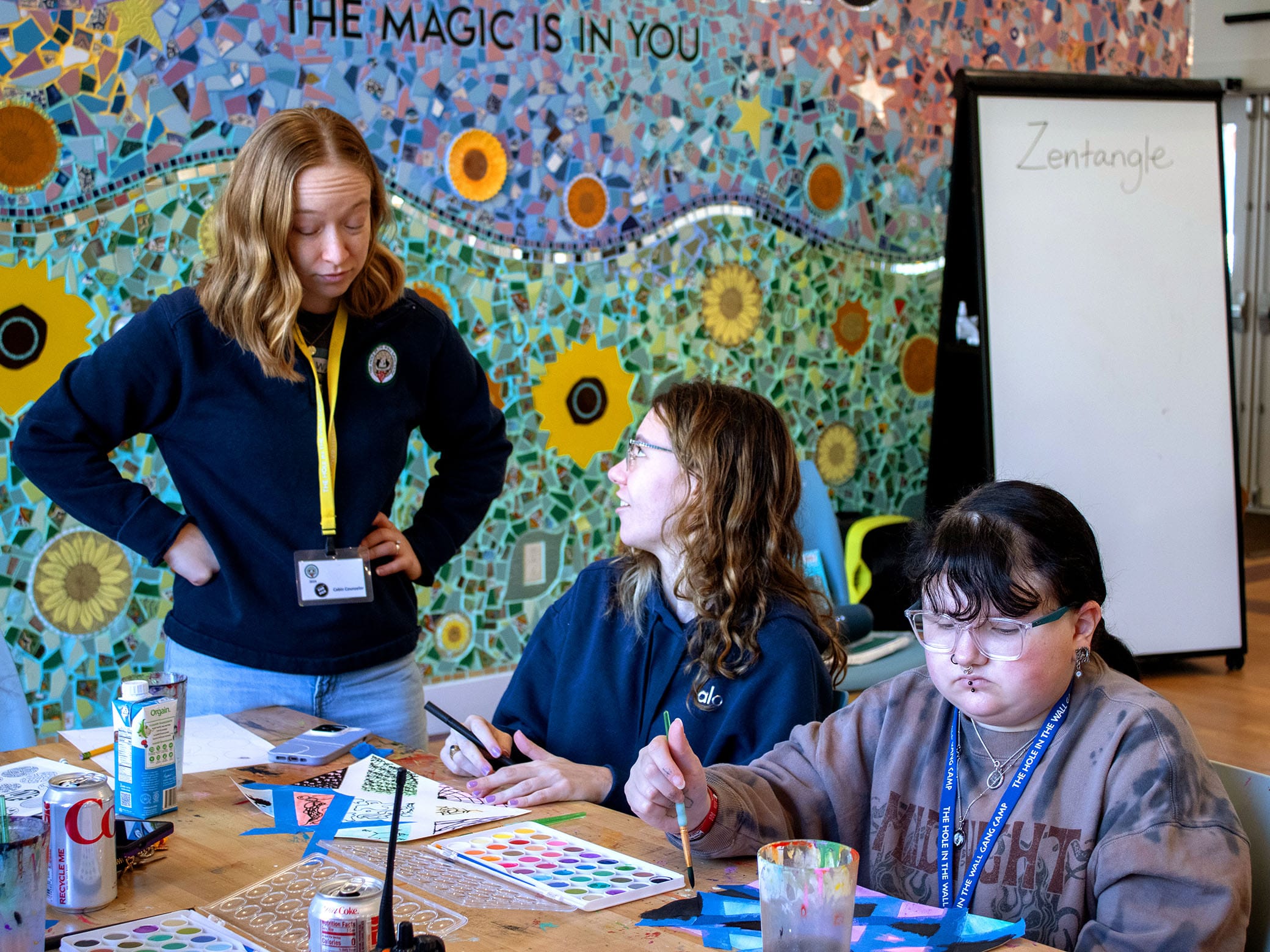 Three people sit at a table painting with watercolors. One woman stands and talks to two others. A colorful mosaic mural is on the wall behind them, and a whiteboard reads Zentangle. Art supplies cover the table.