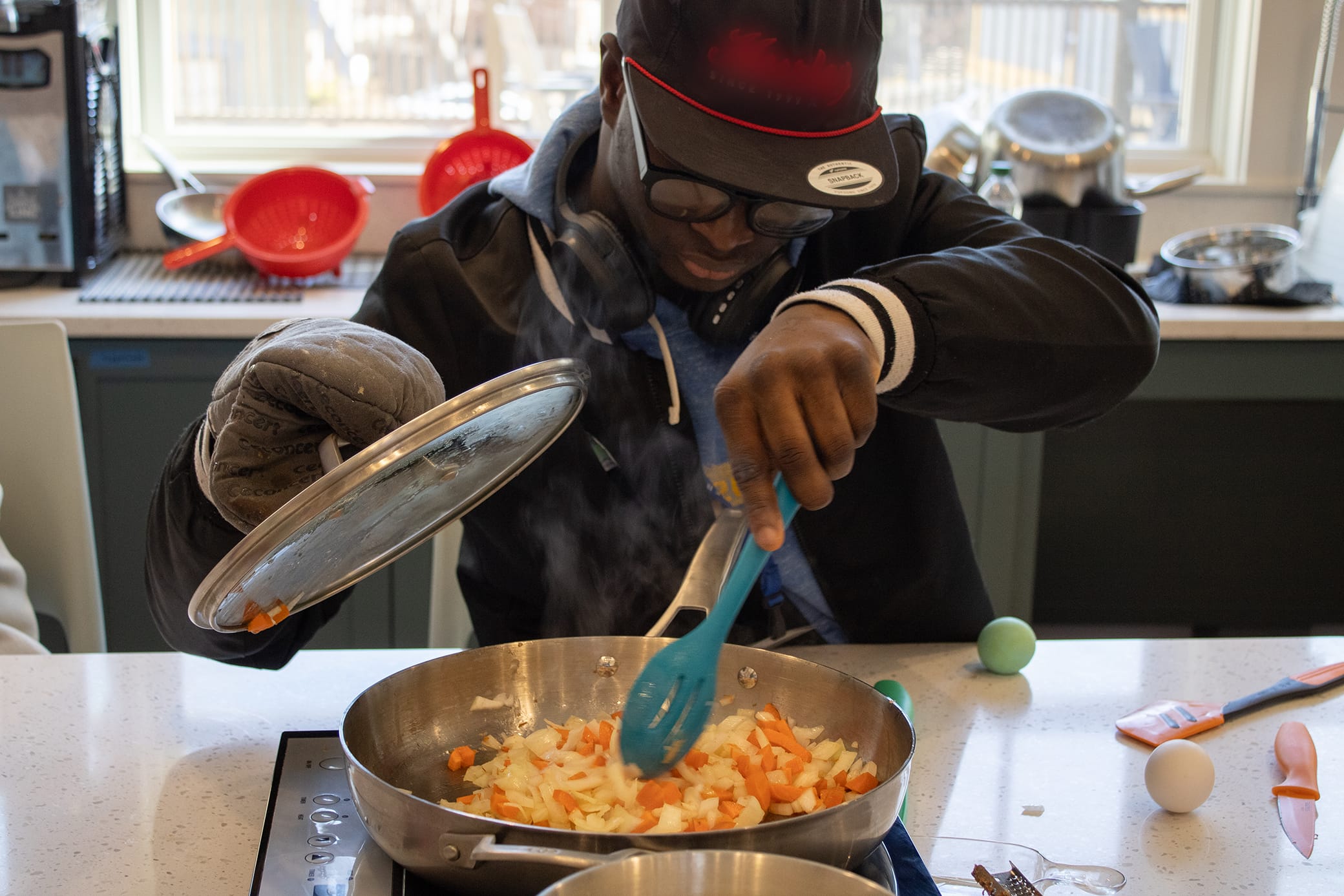 A person wearing glasses and a hat cooks diced vegetables in a pan, lifting the lid with an oven mitt and stirring with a blue spoon. Kitchen tools and ingredients are visible on the counter.