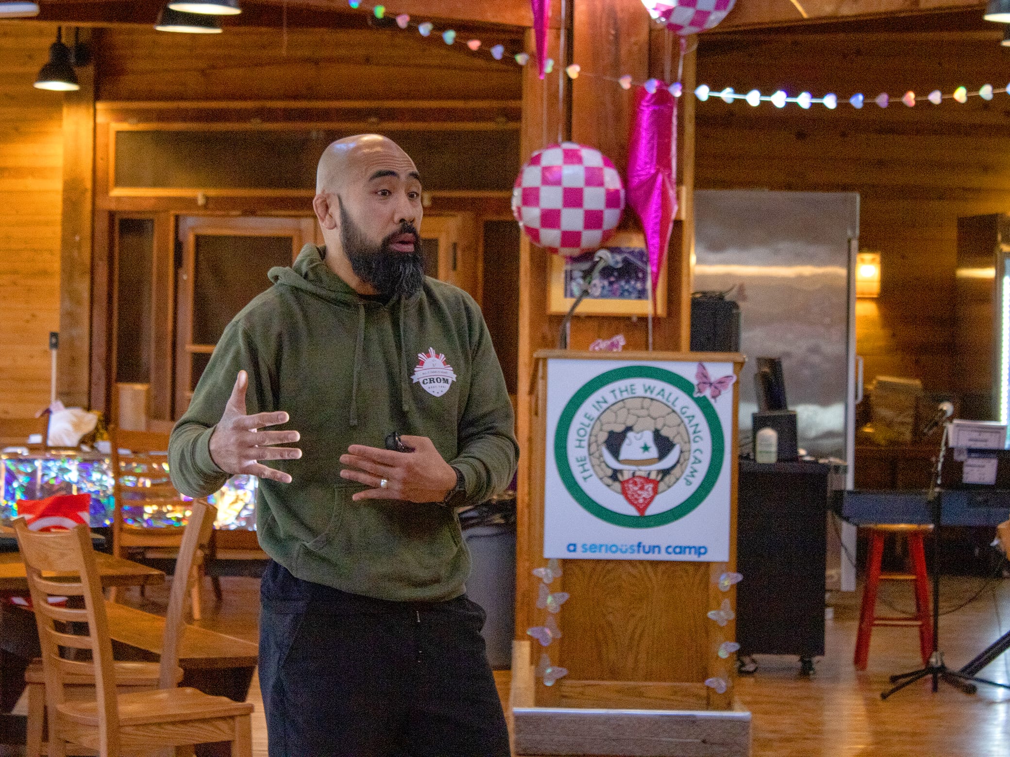 A bearded man in a green hoodie gestures while speaking indoors at a camp facility. Colorful decorations and a sign reading a seriousfun camp are visible in the background.