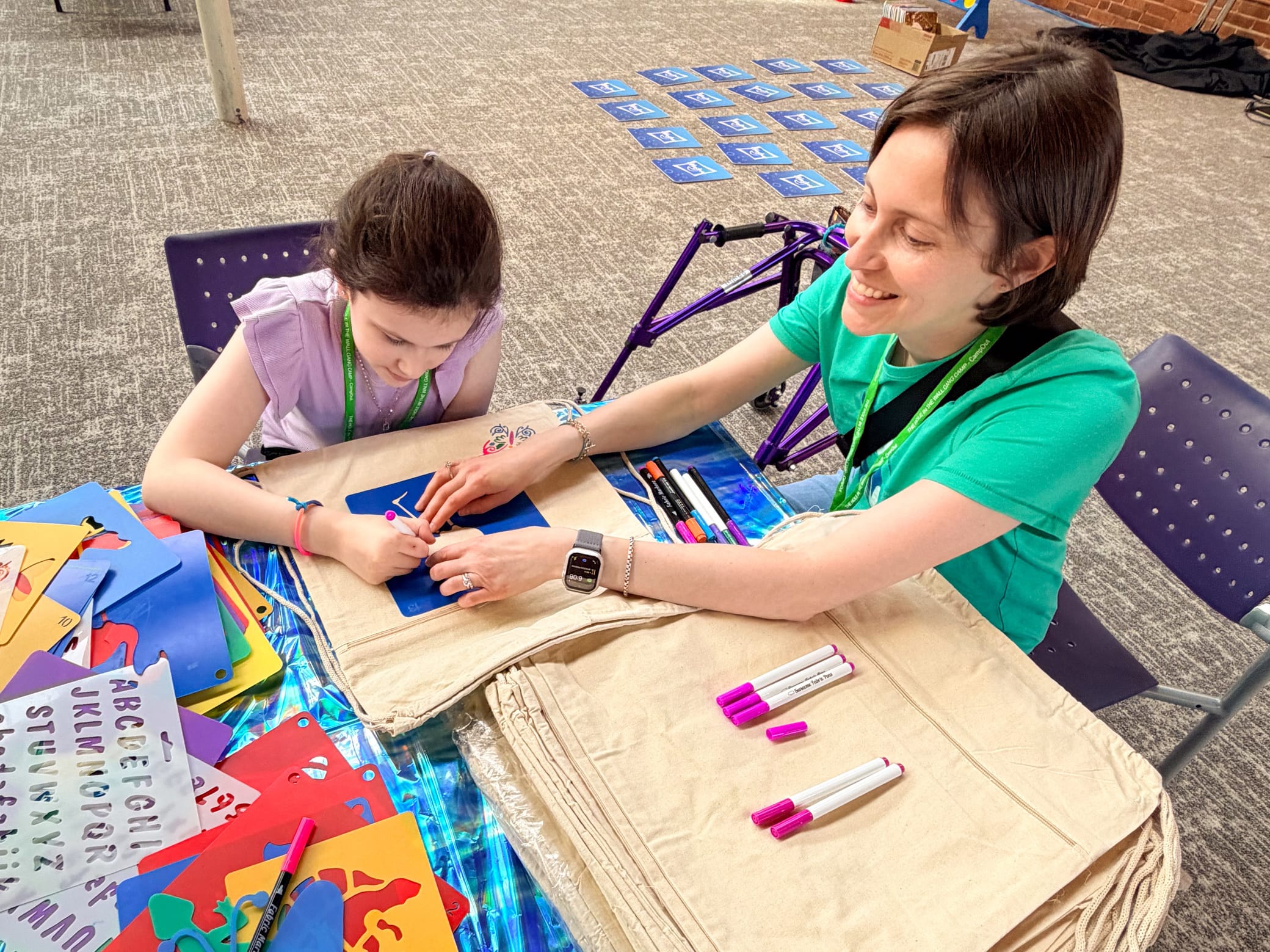 An adult and a child sit at a table, coloring together on cloth bags. Stencils, markers, and craft supplies are spread out around them. Both appear engaged and happy during the art activity.