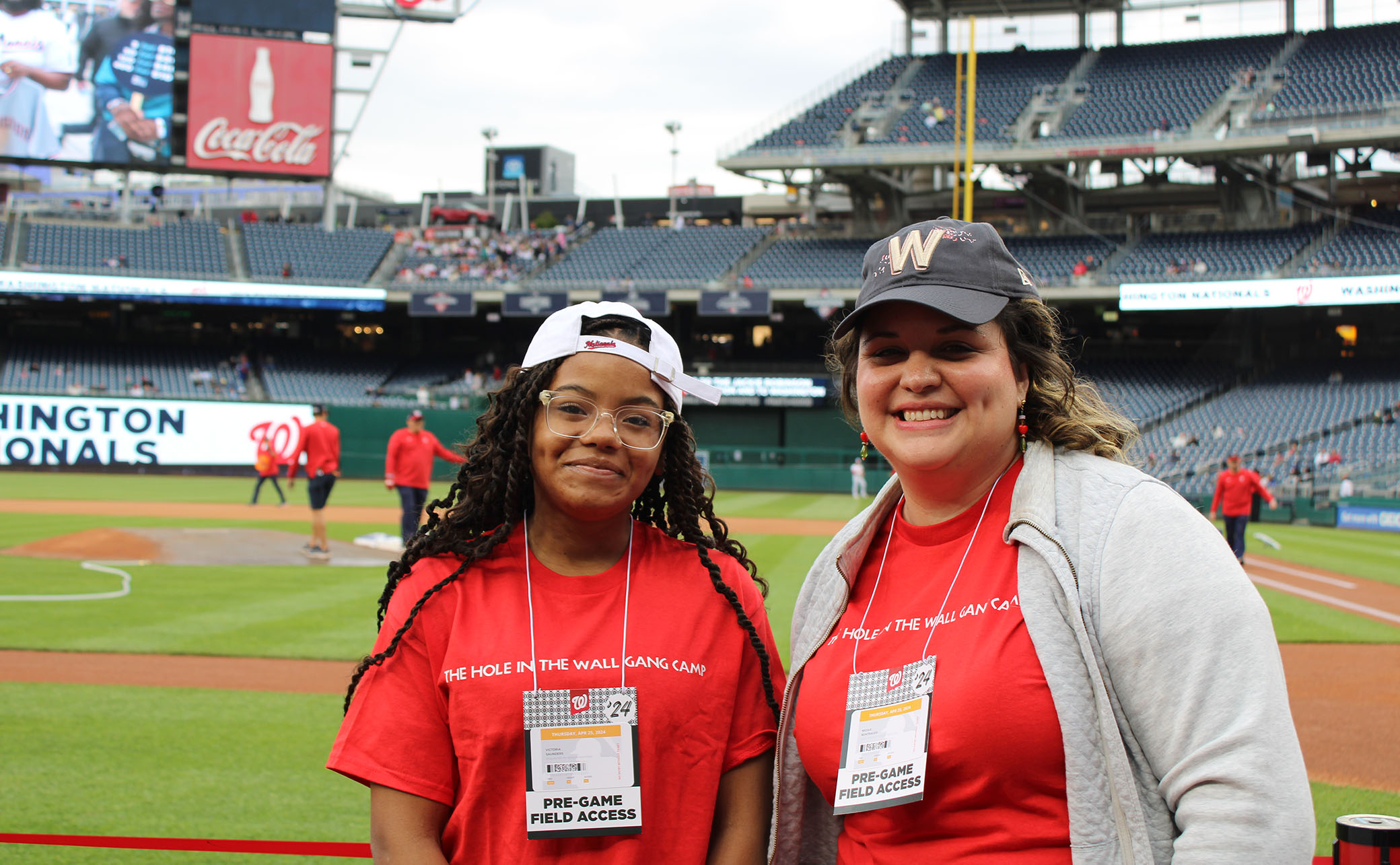 Two smiling women in red shirts and baseball caps stand on a baseball field before a game, wearing badges that say PRE-GAME FIELD ACCESS. The stadium is visible in the background, with players and staff on the field.