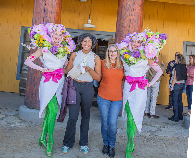 Two smiling women stand between two performers dressed as flowers, with large colorful blossoms on their heads and green leggings. A small group of people stands in the background outside a building with wooden pillars.