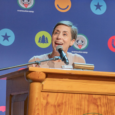 A woman with short hair speaks into a microphone at a wooden podium, smiling. The background features colorful icons, including stars, trees, and smiley faces on a blue backdrop.