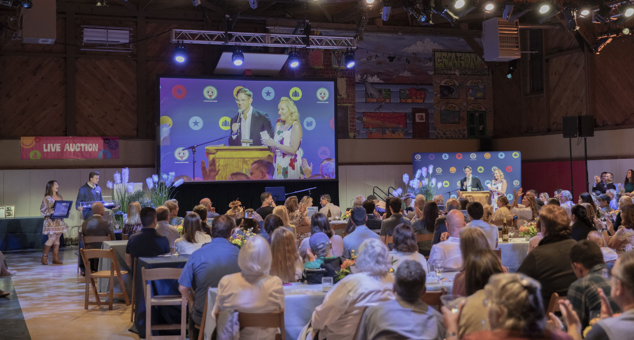 A man and woman speak at a podium on stage during a live auction event, with their image projected on a large screen behind them. Audience members sit at round tables watching.